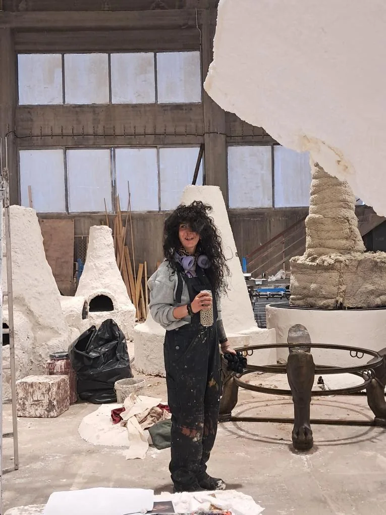 A person in paint-marked workwear standing among large scenic forms and equipment in a workshop.